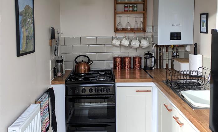 Black gas stove with hob top kettle, boiler, sink and black counter top fridge.Magnetic knife rack on wall. , copper tea, coffee,sugar canisters.